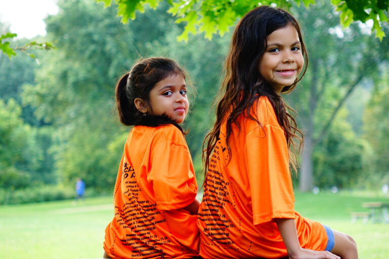 two young girls with long dark brown hair sitting outside. They are facing away from the camera and have their heads turned towards the camera. They are smiling. They are wearing orange shirts with words all over the back for national day for truth and reconciliation.