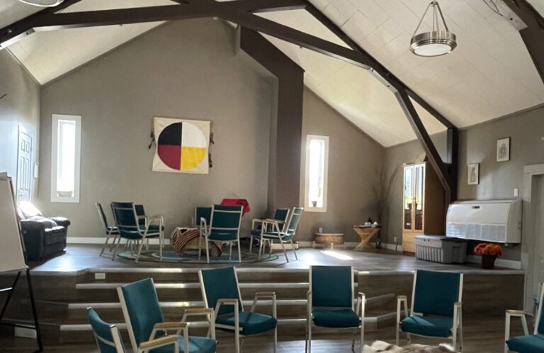 An indoor community gathering space with two circles of chairs around large drums covered with ornate blankets. The space has high ceilings and is low lit by bright sunlight shining through two tall windows on the back wall.
