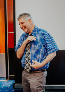 Micheal, a man with dark grey hair, is smiling while facing to his right. He is wearing a blue checkered short sleeved shirt and a tie with various shades of grey and black organized into a grid pattern. He is standing in front of a black projector screen in a room with orange-red wood on the walls. 