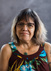 A portrait of a woman with shoulder length dark brown hair that is dyed a light straw colour closer to the ends. She is searing brown a gold rimmed glasses, a gold necklace with a green stone pendant and a brown floral sleeveless top.